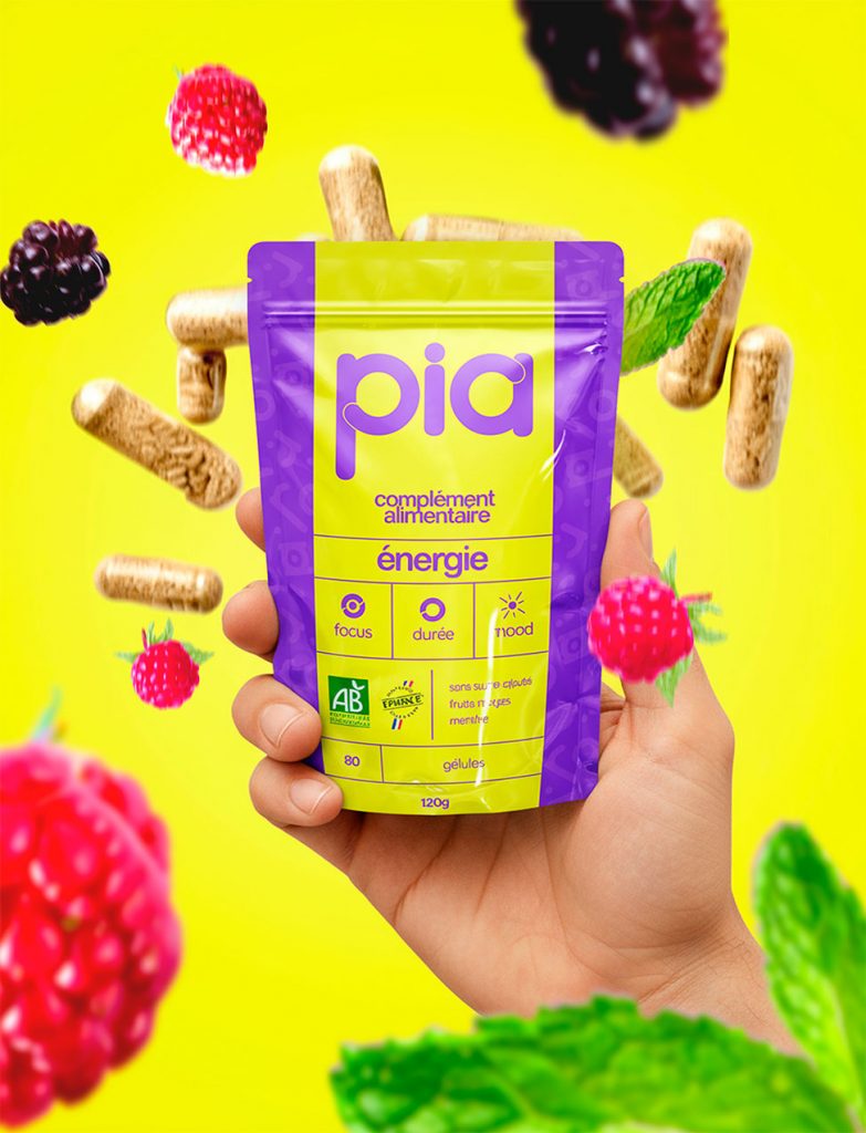 Emballage du complément alimentaire bio PIA Énergie, tenu en main sur fond jaune vibrant, avec gélules naturelles, fruits rouges et feuilles de menthe flottants.